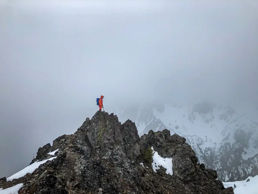 Lone mountaineer standing on a rocky summit in mist above snowfields