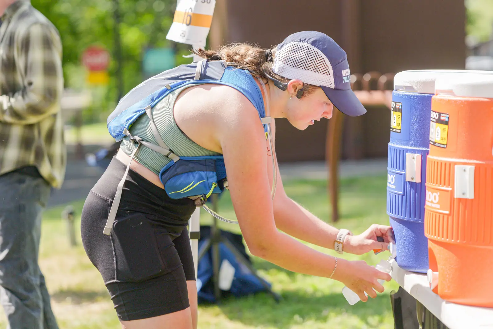 Ultramarathon runner in a hydration vest refilling a bottle at an aid station cooler